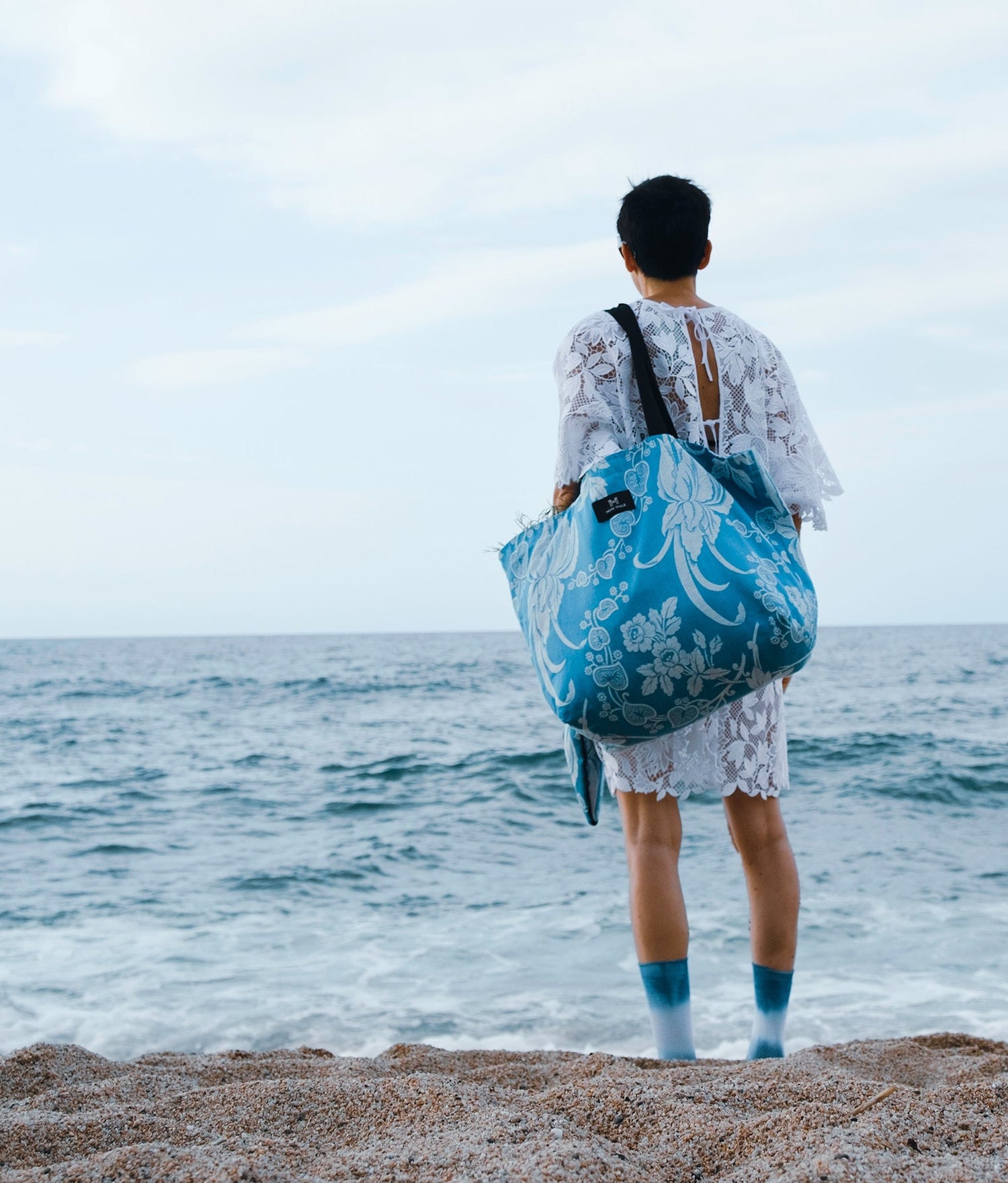 A person standing on a sandy beach facing the ocean, carrying a large blue and white patterned tote bag over their shoulder.