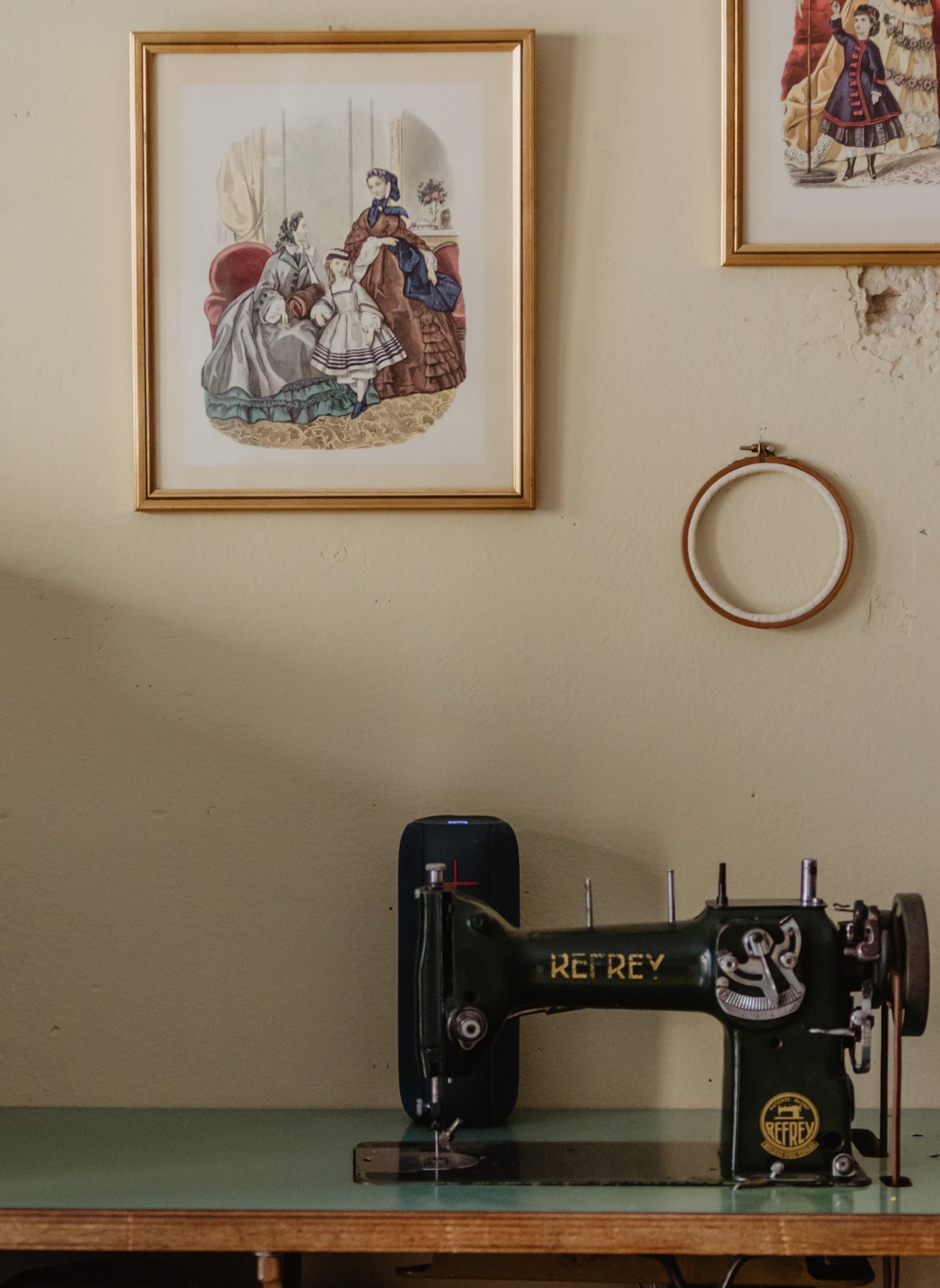A vintage black Metabole sewing machine sits on a green-tinted wooden table against a beige wall decorated with framed illustrations, a wooden hoop, and keys. To the left is a wooden chest of drawers.