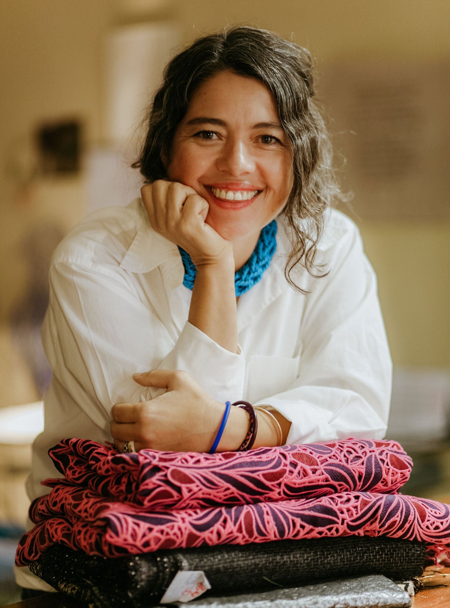 A smiling woman with dark hair sits behind a stack of folded patterned fabrics, wearing a white shirt and a bright blue necklace in a warm, softly lit indoor setting.