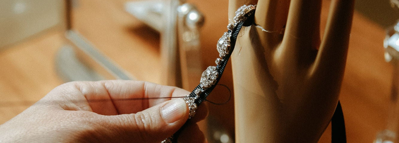 A hand holds a silver-toned beaded bracelet with a black cord, shown next to a beige jewelry display mannequin hand on a wooden table.