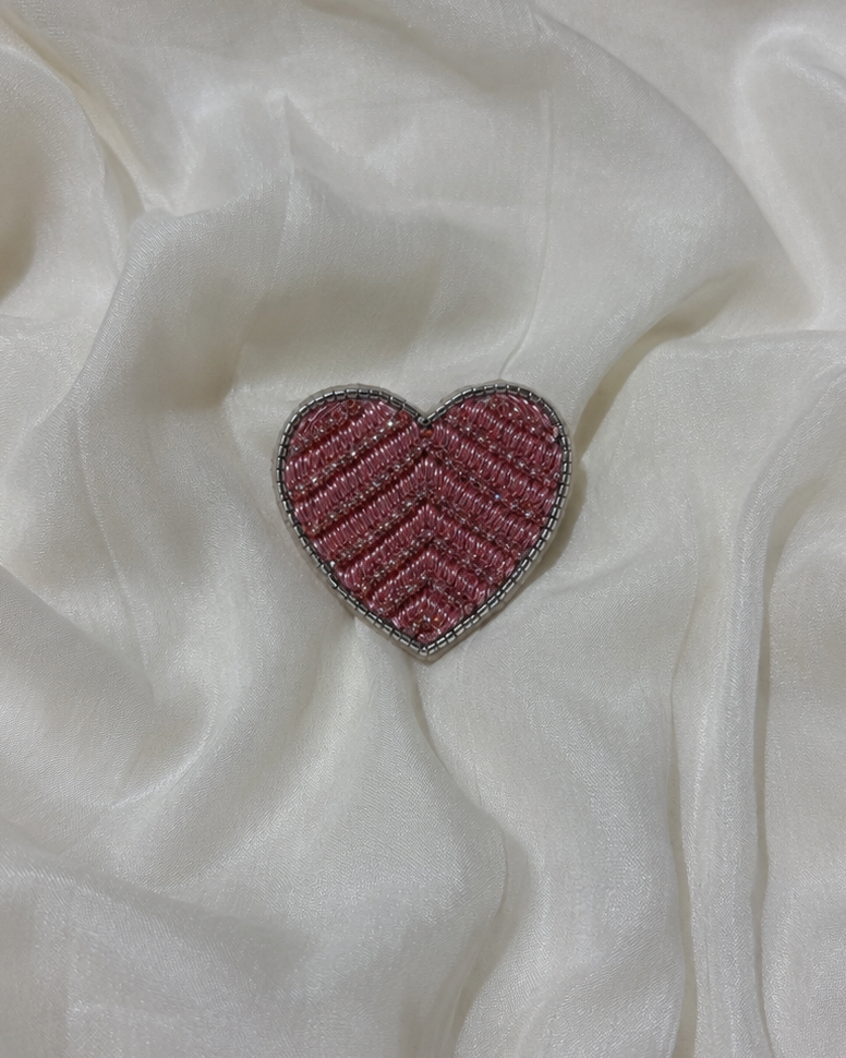A small heart-shaped brooch with a pink and silver chevron pattern and rhinestone border, resting on draped white fabric.